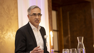 Mann mit grau-meliertem Haar und Brille spricht stehend am Podium mit einem Headset-Mikrofon. Den Hintergrund ziert eine beige-gemusterte Wand. Ebenfalls ersichtlich sind eine Karaffe Wasser und drei Gläser.