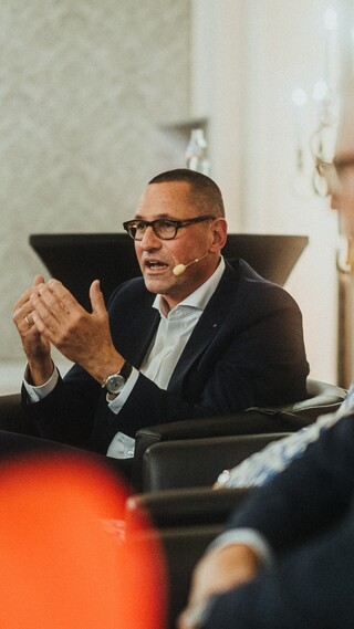 Ein sitzender Herr in Anzug mit Headset spricht und gestikuliert.
