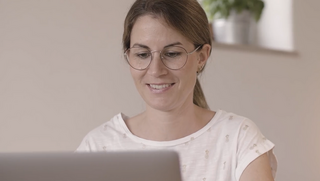 Frau mit Brille, freundlichem Ausdruck und weißem T-Shirt schaut in Laptop.