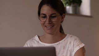Frau mit Brille, freundlichem Ausdruck und weißem T-Shirt schaut in Laptop.