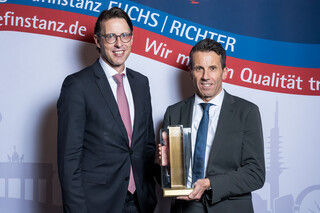 Zwei lächelnde Herren in Anzug posieren mit einer Trophäe vor einer Fotowand.