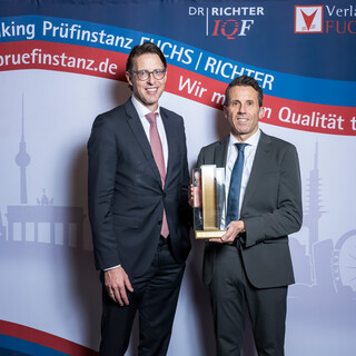 Zwei lächelnde Herren in Anzug posieren mit einer Trophäe vor einer Fotowand.
