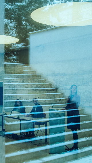 Zwei sitzende Personen und eine stehende Dame sind hinter einer Glasfront zu sehen. In Glas spiegelt sich ein Treppenaufgang im Außenbereich.