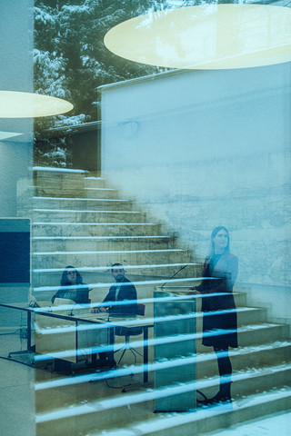 Zwei sitzende Personen und eine stehende Dame sind hinter einer Glasfront zu sehen. In Glas spiegelt sich ein Treppenaufgang im Außenbereich.