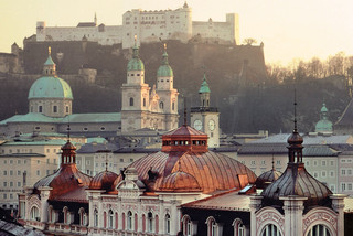 Stammhaus des Bankhaus Spängler in der Schwarzstraße 1 in 5020 Salzburg von oben. Barocke Kuppeln und eine dunkelrosa Fassade sind ersichtlich. Im Hintergrund sieht man die Festung Hohensalzburg und die Domkuppeln.