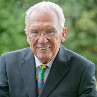 Älterer Mann mit grauem Harr und Brille, der freundlich lächelt. Er trägt ein dunkelgraues Sakko mit weißem Hemd und bunter Krawatte.