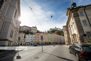 Straßenzufahrt in der Stadt Salzburg mit mehreren historischen Gebäuden, welche direkt aneinander gebaut sind. Sie haben unterschiedliche pastelltönige Farben (orange, blau, grün, rosa). Im Hintergrund leicht erkennbar ist die Festung Hohensalzburg.