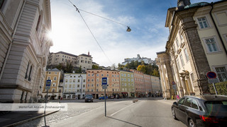 Straßenzufahrt in der Stadt Salzburg mit mehreren historischen Gebäuden, welche direkt aneinander gebaut sind. Sie haben unterschiedliche pastelltönige Farben (orange, blau, grün, rosa). Im Hintergrund leicht erkennbar ist die Festung Hohensalzburg.