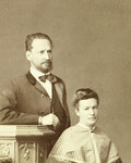 Historische Bild in beiger Farbe, das eine Frau (sitzend) und einen stehenden Mann hinter ihr zeigt. Vermutlich sind sie ein Ehepaar. Der Mann trägt ein Frack und eine Fliege, er hat gekrauste Haare und einen Bart. Die Frau trägt eine festlichen Umhang und hat die Haare hochgestockt.