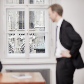 2 Männer, einer davon stehend, der andere sitzend, unterhalten sich. Der Blick aus dem Fenster im Hintergrund ist scharf und die Fassade des Stephansdom Wien ersichtlich. 