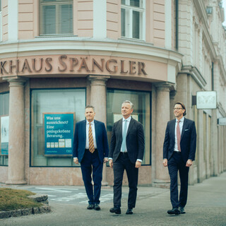 Drei Männer in Anzügen gehen bei nebligem Wetter vor dem historischen Bankhaus Spängler nach vorne.