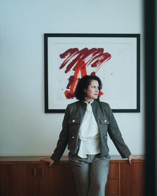 Eine Dame in Geschäftskleidung stützt sich mit den Händen auf eine Kommode und blickt zur Seite.