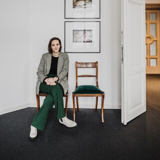 Eine junge Frau sitzt in einem modernen Anzug auf einem Stuhl. An der Wand hängen zwei Bilder, und rechts ist eine offene Tür sichtbar.