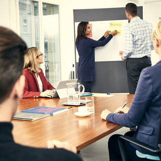 Zwei Männer und drei Frauen unterhalten sich in einem hellen Seminarraum. Am Tisch liegen ein Broschüre und Stifte, ein Flipchart ist im Hintergrund. Dort stehen ein Mann und eine Frau.