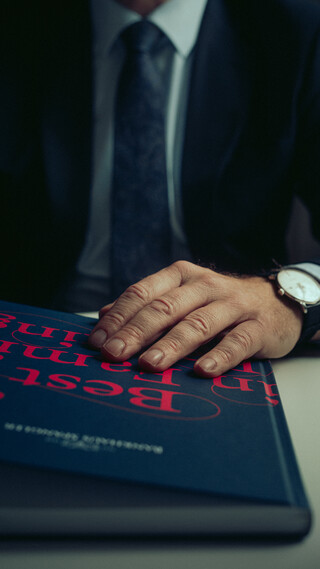 Eine Detailaufnahme zeigt eine Broschüre in den Händen eines Mannes; an seinem Handgelenk trägt er eine Uhr.