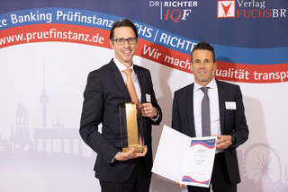 Zwei Männer in schwarzen Anzügen, weißen Hemden und Krawatten stehen vor einer Wand mit der Aufschrift "Beste Banking Prüfinstanz Fuchs, Richter und www.pruefinstanz.de". Ein Mann hält eine Urkunde in Händen, der andere eine Art Trophäe.