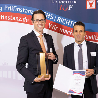 Zwei Männer in schwarzen Anzügen, weißen Hemden und Krawatten stehen vor einer Wand mit der Aufschrift "Beste Banking Prüfinstanz Fuchs, Richter und www.pruefinstanz.de". Ein Mann hält eine Urkunde in Händen, der andere eine Art Trophäe.