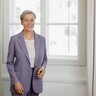 Eine Dame steht vor dem Fenster in einem Blazer und lächel