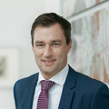 Mann mit braunem Haar lächelt freundlich. Er trägt ein weißes Hemd, ein dunkelblaues Sakko sowie eine rot-blau-weiß-gemusterte Krawatte.