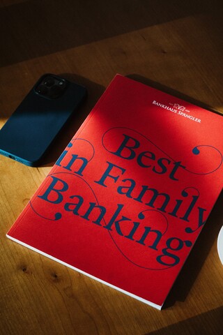 Eine Broschüre mit dem Titel "Best in Family Banking" liegt auf einem Tisch neben einem Smartphone.