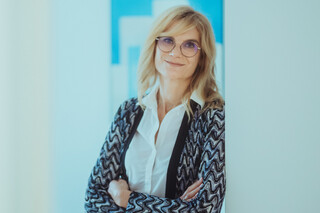 Eine blonde Frau mit weißer Bluse, Brille und bunter Jacke lehnt am Türrahmen. Im Hintergrund hängt ein Bild an der Wand.
