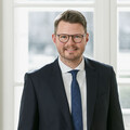Ein lächelnder Herr in Anzug steht hinter einem leeren Besprechungstisch.