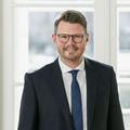 Ein lächelnder Herr in Anzug steht hinter einem leeren Besprechungstisch.