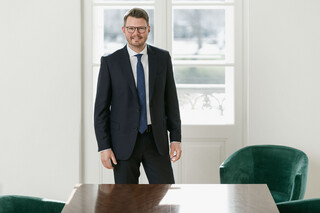Ein lächelnder Herr in Anzug steht hinter einem leeren Besprechungstisch.