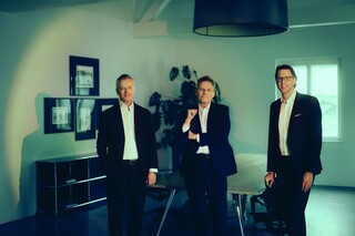 Drei Personen in Business-Kleidung stehen in einem Besprechungsraum und blicken in die Kamera.