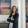 Ein Beraterin lehnt am Türrahmen in einem Bürogebäude des Bankhaus Spängler. Sie lächelt freundlich, im Hintergrund sind ein buntes Bild und ein Schrank ersichtlich. 