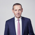 Dr. Markus Ploner Ein Mann in blauem Anzug mit rosa Krawatte steht vor einer grauen Wand.