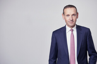 Ein Mann in blauem Anzug mit rosa Krawatte steht vor einer grauen Wand.