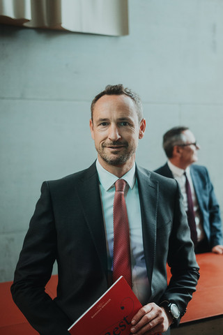 Ein lächelnder Herr in Anzug hält eine Broschüre in der Hand.