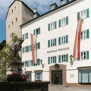 Fassade der Niederlassung Zell am See des Bankhaus Spängler. Historisches Gebäude mit grünen Fensterbalken und zwei Fahnen in rot und weiß (1x Österreich-Wappen, 1x Salzburg-Wappen), der Stadtbrunnen ist ebenfalls am Bild.