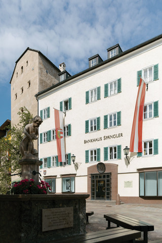 Fassade der Niederlassung Zell am See des Bankhaus Spängler. Historisches Gebäude mit grünen Fensterbalken und zwei Fahnen in rot und weiß (1x Österreich-Wappen, 1x Salzburg-Wappen), der Stadtbrunnen ist ebenfalls am Bild.