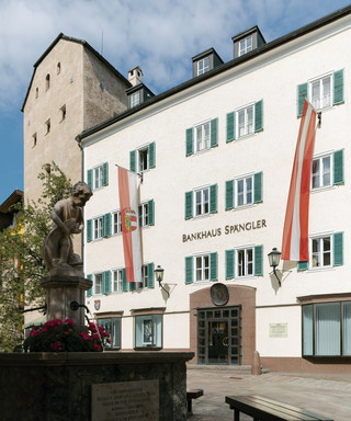 Fassade der Niederlassung Zell am See des Bankhaus Spängler. Historisches Gebäude mit grünen Fensterbalken und zwei Fahnen in rot und weiß (1x Österreich-Wappen, 1x Salzburg-Wappen), der Stadtbrunnen ist ebenfalls am Bild.