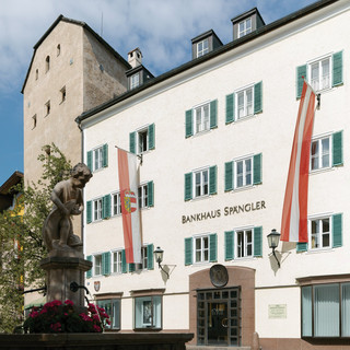 Fassade der Niederlassung Zell am See des Bankhaus Spängler. Historisches Gebäude mit grünen Fensterbalken und zwei Fahnen in rot und weiß (1x Österreich-Wappen, 1x Salzburg-Wappen), der Stadtbrunnen ist ebenfalls am Bild.