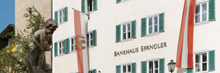 Fassade der Niederlassung Zell am See des Bankhaus Spängler. Historisches Gebäude mit grünen Fensterbalken und zwei Fahnen in rot und weiß (1x Österreich-Wappen, 1x Salzburg-Wappen), der Stadtbrunnen ist ebenfalls am Bild.
