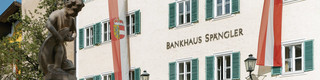 Fassade der Niederlassung Zell am See des Bankhaus Spängler. Historisches Gebäude mit grünen Fensterbalken und zwei Fahnen in rot und weiß (1x Österreich-Wappen, 1x Salzburg-Wappen), der Stadtbrunnen ist ebenfalls am Bild.