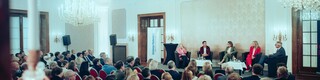 Fünf Sprecher sitzen vor einem Publikum in einem großen Saal, der mit zwei Kronleuchtern ausgestattet ist.