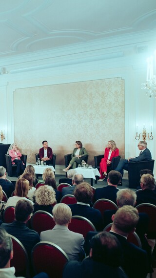 Fünf Sprecher sitzen vor einem Publikum in einem großen Saal, der mit zwei Kronleuchtern ausgestattet ist.