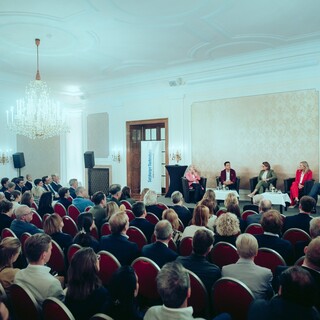 Fünf Sprecher sitzen vor einem Publikum in einem großen Saal, der mit zwei Kronleuchtern ausgestattet ist.