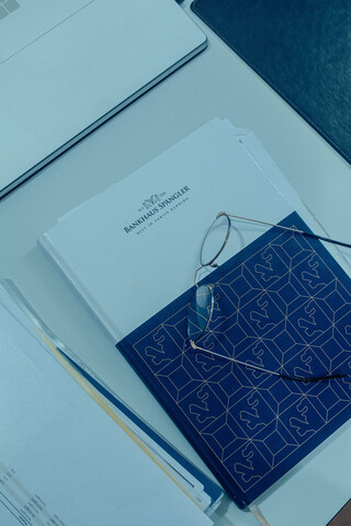 Auf dem Tisch liegt ein Notizbuch mit dem Logo vom Bankhaus Spängler. Darauf befindet sich eine Brille, links davon liegen Unterlagen mit Zahlen.