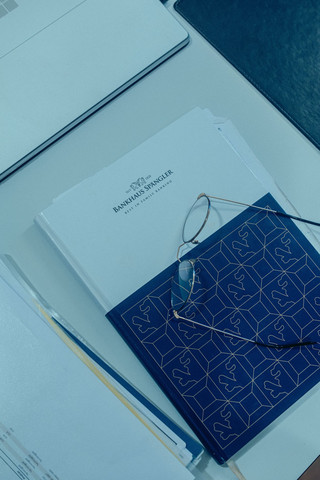 Auf dem Tisch liegt ein Notizbuch mit dem Logo vom Bankhaus Spängler. Darauf befindet sich eine Brille, links davon liegen Unterlagen mit Zahlen.
