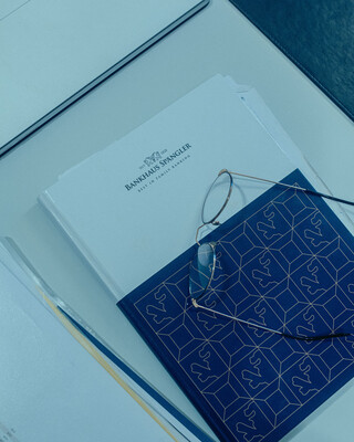 Auf dem Tisch liegt ein Notizbuch mit dem Logo vom Bankhaus Spängler. Darauf befindet sich eine Brille, links davon liegen Unterlagen mit Zahlen.