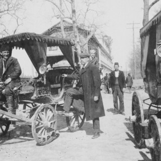 Historische Schwarz-Weiß-Aufnahme einer Straßenszene mit mehreren Personen und Pferdewagen.