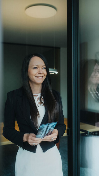 Person in Business-Kleidung hält eine Broschüre in den Händen und steht in einem modernen Bürobereich.