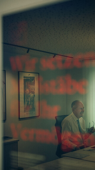 Ein Mann sitzt an seinem Schreibtisch – im Hintergrund spiegelt sich dezent unser Leitgedanke: „Wir setzen Maßstäbe für Ihr Vermögen.