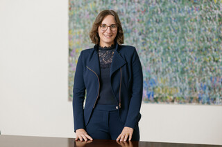 Eine Frau mit braunen Haaren und Brille steht in blauer Jacke und Bluse an einem Tisch vor einem großen Bild.