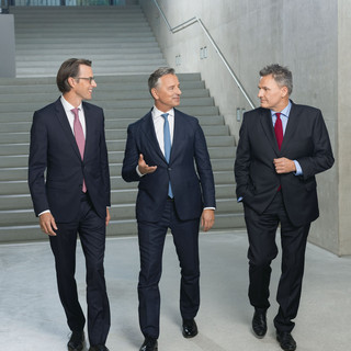 Drei Männer in schwarzen Anzügen vor einer Treppe in modernem Stil, sie beraten sich im Gehen. Am Ende der Treppe ist eine Glastür.
